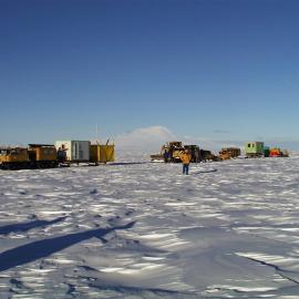 Person on skidoo in front of person on sled on textured snow with scientific equipment on sled.  Green portacabin in background